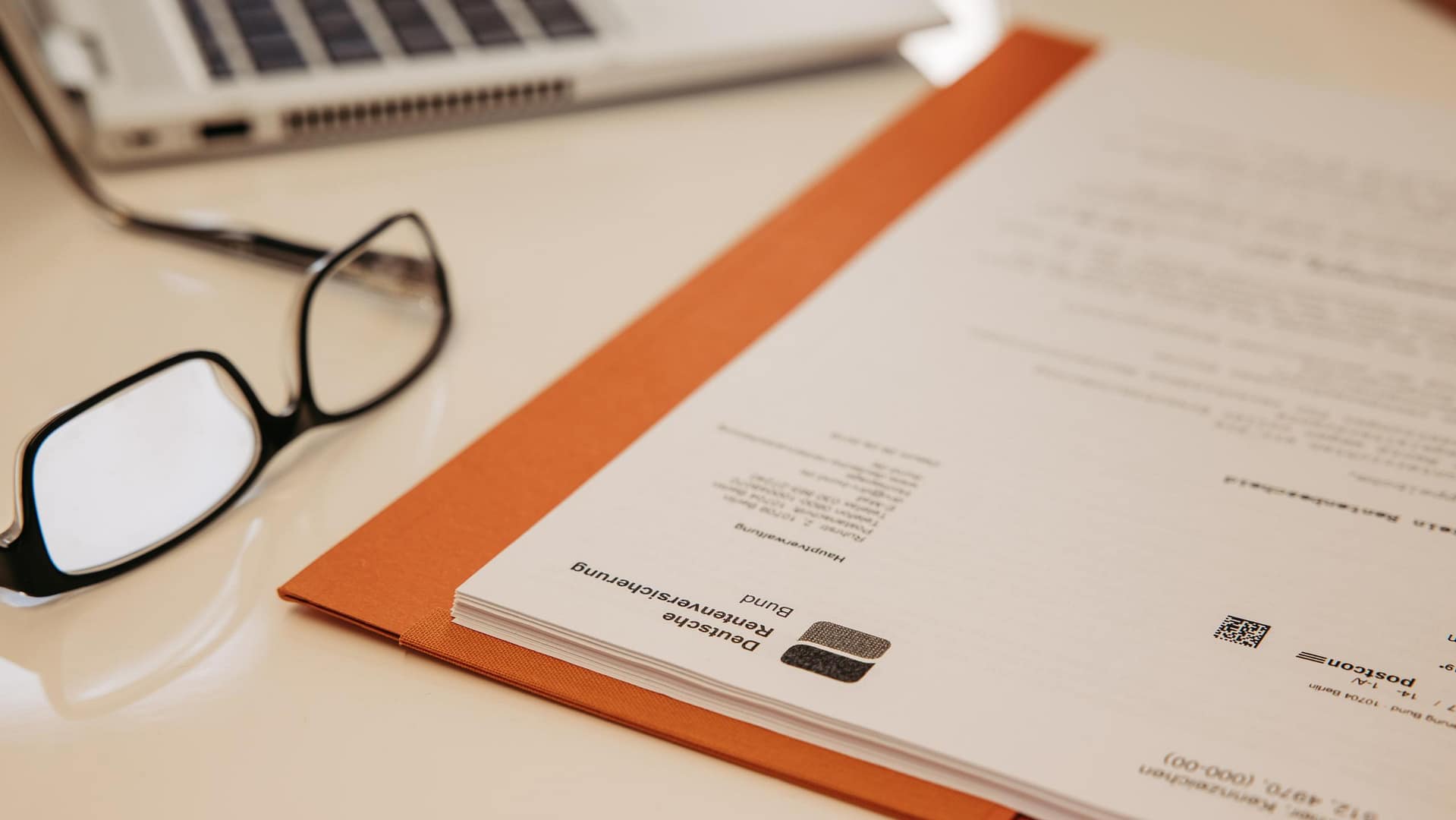 Unterlagen zur Rentenberatung – Kanzlei van Heiden Professionelle Rentenberatung: Dokumente der Deutschen Rentenversicherung Bund in einer orangen Akte auf einem Schreibtisch mit Lesebrille und Laptop.